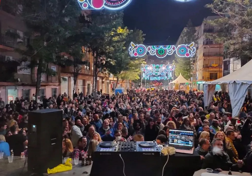 Plaza llena de gente de noche, con la cabina del DJ delante y las luces de fiesta al fondo