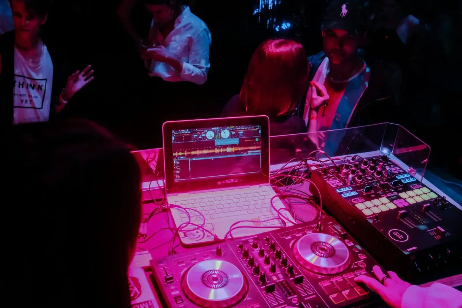 Mesa de mezclas y portátil de un DJ iluminados en rosa, con gente bailando detrás