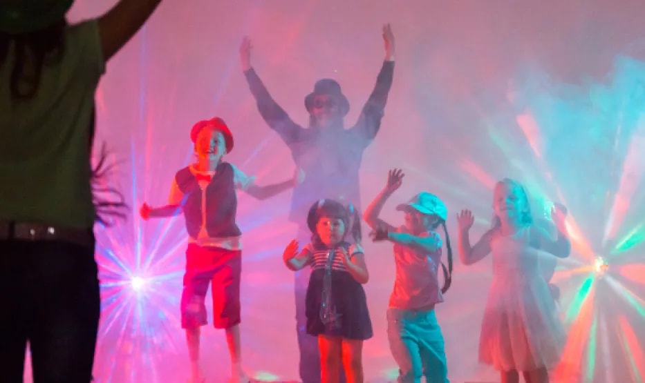 Cuatro niños bailando con los brazos en alto entre focos de colores y humo